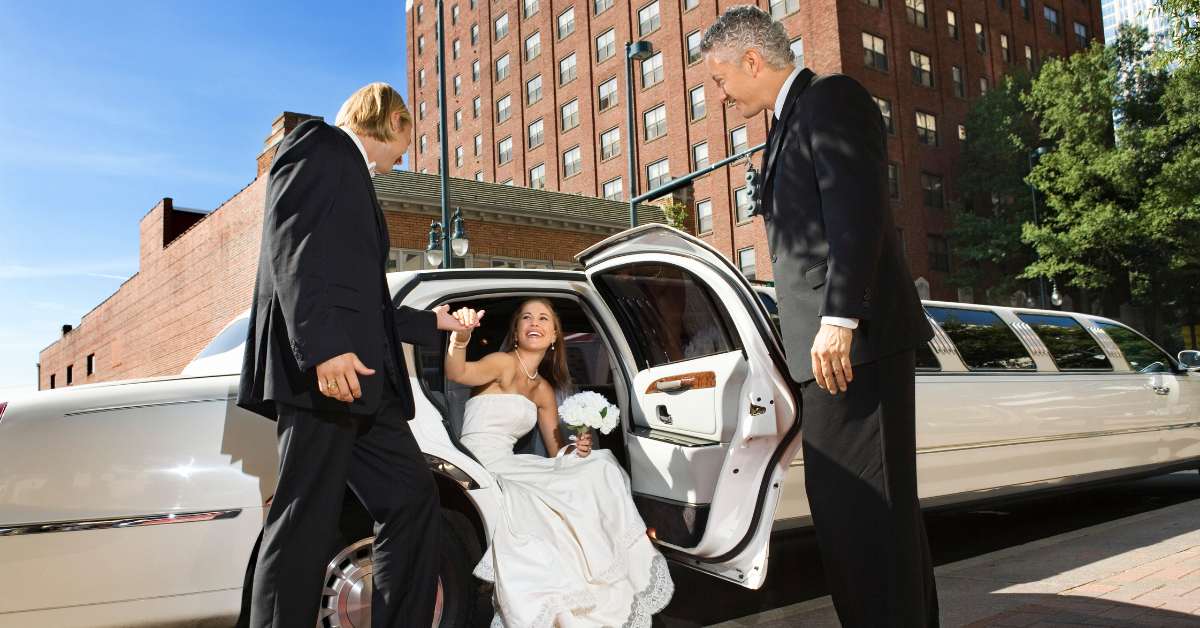 Bride leaving limousine on a bright day, assisted by two men in suits