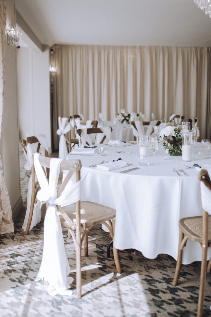 Elegantly decorated banquet hall with round tables set with crisp white linens, fine glassware, and floral centerpieces, illuminated by warm ambient lighting, creating a sophisticated and inviting atmosphere